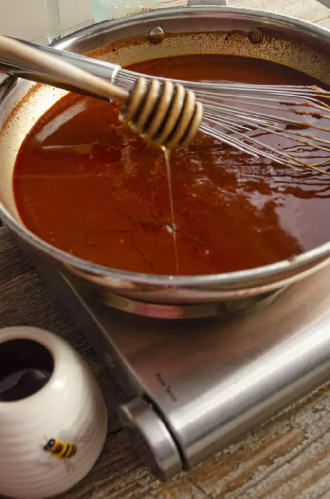 Honey drips from a honey wand down into a stainless steel skillet filled with a freshly cooked batch of red chile sauce.