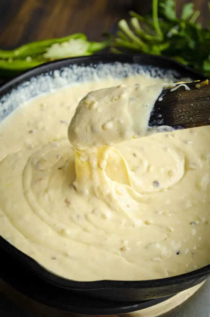 Light yellow, and melty; Green Chile Queso is lifted up with a spoon and drips back down into a cast iron skillet.