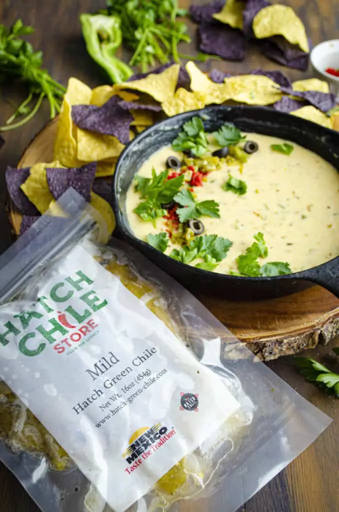 A black cast iron skillet is full of light yellow melted queso cheese, garnished with chopped chile, sliced olives and cilantro leaves. The skillet is surrounded by blue and yellow corn tortilla chips and a bag of frozen Hatch Chile Store chile is placed next to it all.