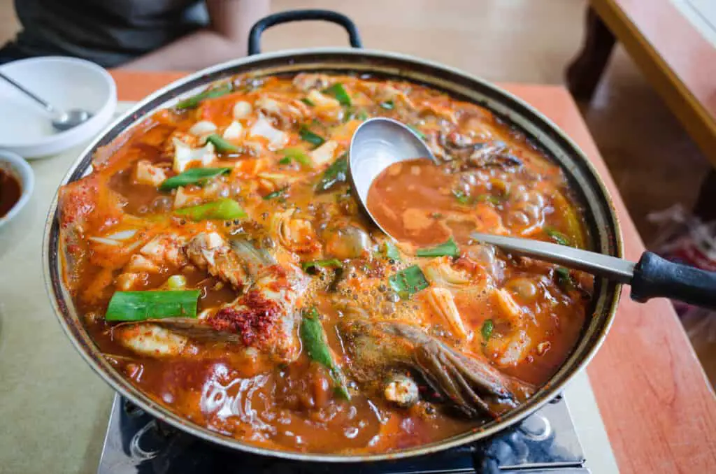 A large pot of thick stew or gumbo boils, full of colorful and varied ingredients. A metal ladle has been left to rest in the center and is partially submerged in the stew.