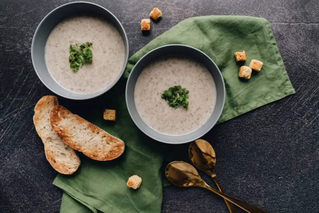 Looking down on two bowls of creamy pureed mushroom soup in grey bowls on a dark grey surface. Each bowl is garnished with springs of parsley in the center. Crusty bread and gold spoons lie adjacent to the bowls.