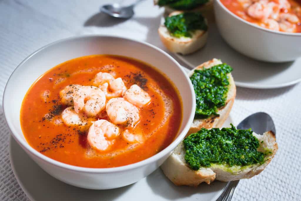 A white bowl an a white tablecloth holds a bright orange red serving of smooth and creamy shrimp bisque with several cooked shrimp pieces in the center. Two slices of crusty bread, spread with green pesto, sit adjacent.