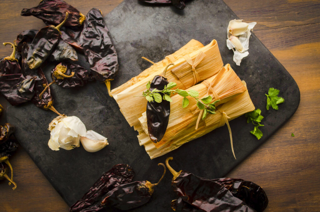 Looking down on a stack of tamales still in their corn husks, surrounded by dried red chile pods.