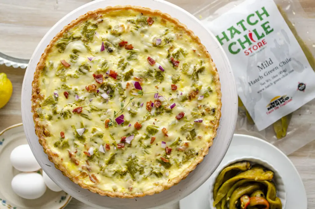 Looking down onto a Green Chile bacon quiche garnished with red onion and a sprig of thyme. Displayed next to it are some eggs, whole roasted and peeled green chiles, and a packet of Mild Hatch Green Chile from The Hatch Chile Store.