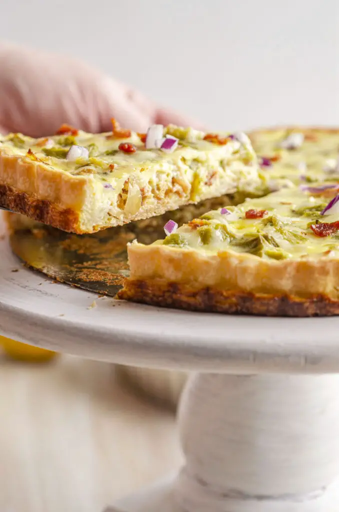 A white wooden cake stand displays a Green Chile Bacon Quiche with a slice being removed.