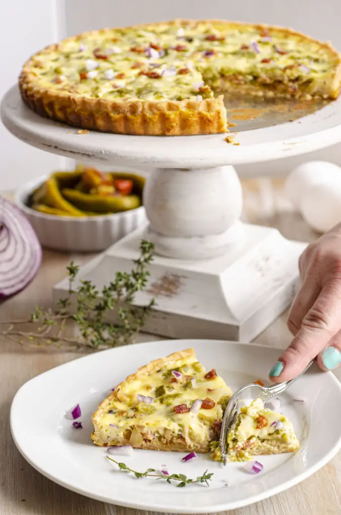 A quiche is displayed on a white wooden cake stand in the background, along with a slice of quiche on a white plate in the foreground.