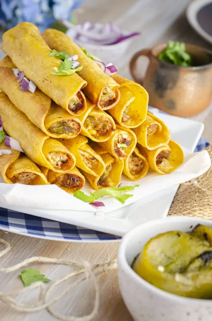 Looking into golden fried Taquitos so the viewer can see they are stuffed with green chile, chicken, cream cheese, salsa, and cheese stacked in a pyramid shape on white and blue plates. A garnish of minced red onions and green cilantro leaves are sprinkled over the top.