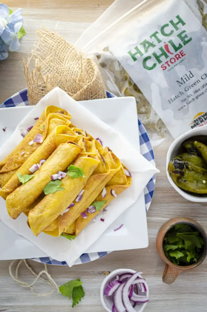 Looking straight down from above onto a stack of golden fried green chile chicken taquitos surrounded by bowls filled with sliced red onion, cilantro leaves, whole roasted green chiles, and a Hatch Chile Store freezer pouch filled with whole green chiles.