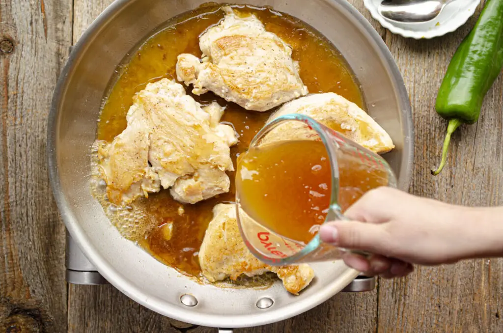 Pouring an orange glaze mixture into sauteed chicken breast in a skillet.