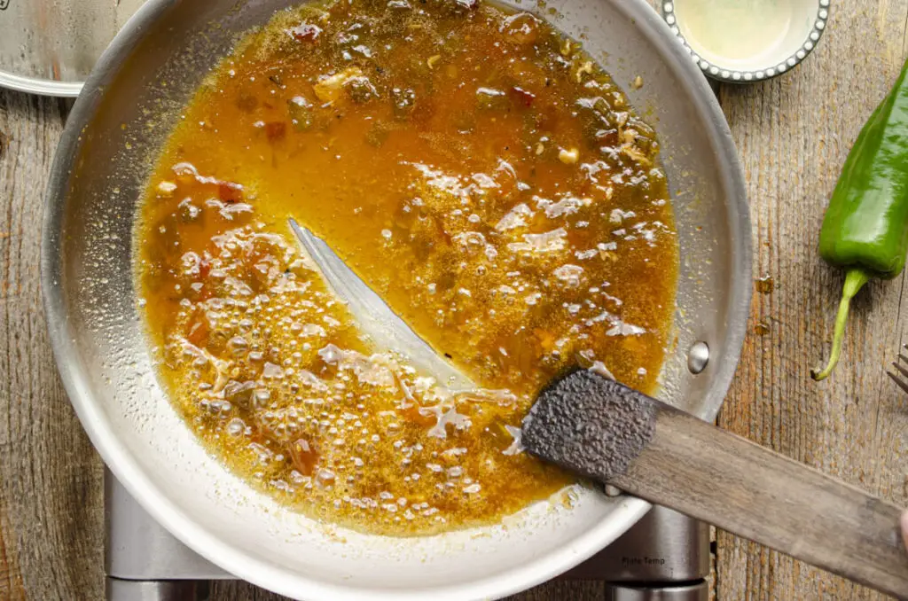 A wooden cooking utensil is pulled across the bottom of a skillet that is cooking a Green Chile Glaze, leaving a trail in the glaze.
