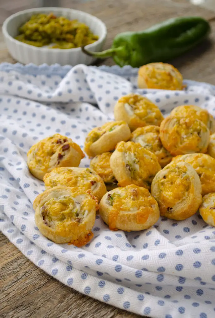 Several bacon green chile puffy pinwheels placed on a blue and white polkadot kitchen towel. A bowl of chopped green chile and a whole Hatch green chile can be seen in the background.