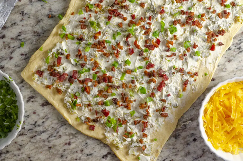 Looking down on a granite counter where a rectangle of dough has been spread with cream cheese, green chile, cooked bacon bits and chopped scallions.