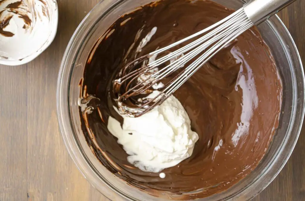 A glass mixingbowl contains melted shiny chocolate with a dollop of sour cream being mixed in by a whisk to make the Johnny Depp Trial Muffins Recipe