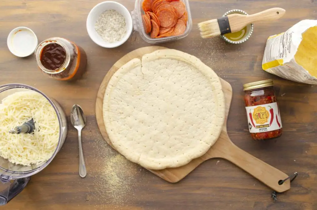 Looking down onto a pizza work station with a pizza crust on a peel, surrounded by ingredients and sauces.