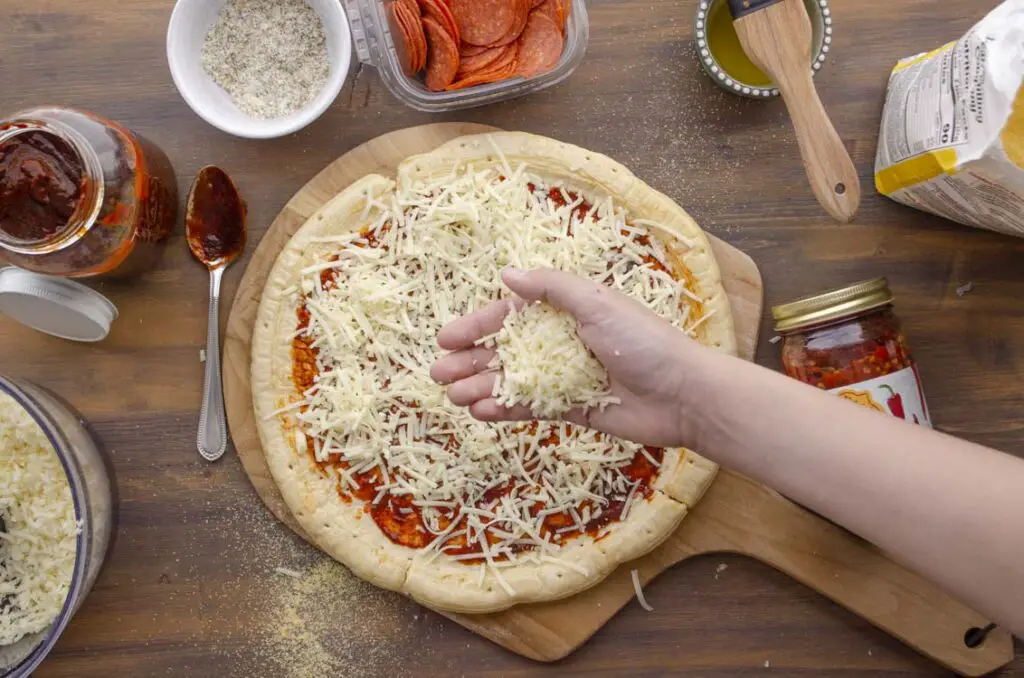 A hand is shown spreading shredded mozzarella onto pizza dough.