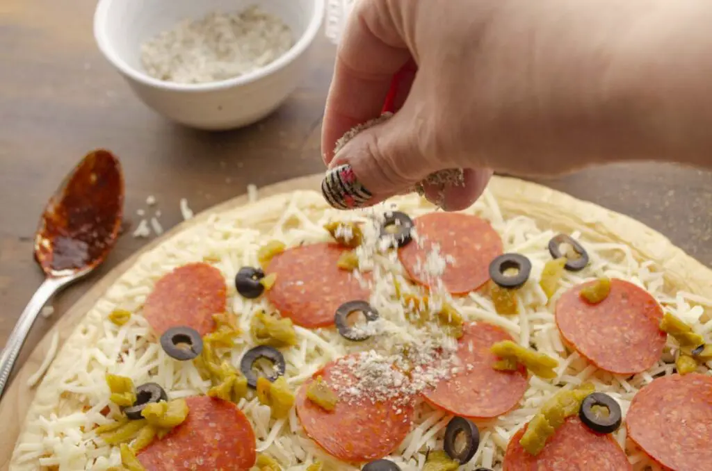 A hand is sprinkling seasonings onto a freshly prepared pizza.