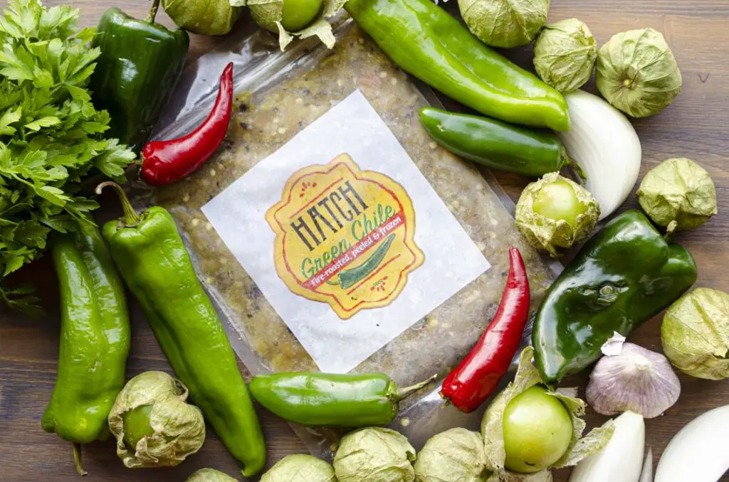 A Hatch green chile pouch surrounded by fresh green and red chiles, sliced white onion garlic cloves and tomatillos.