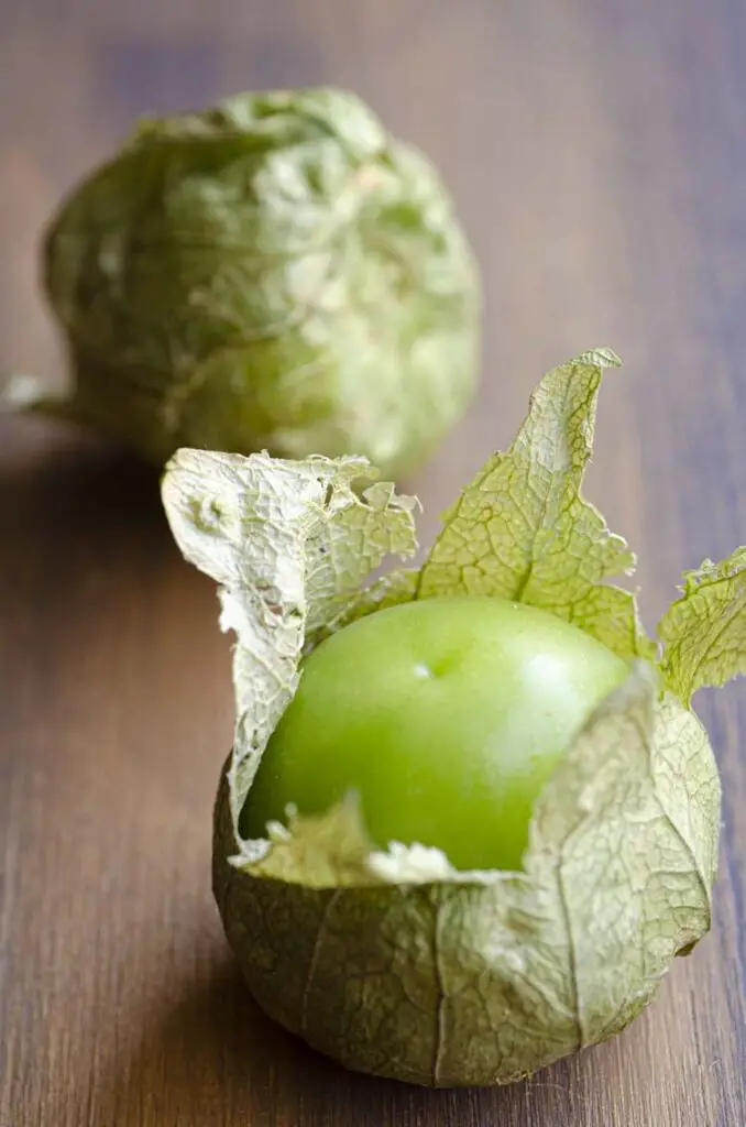 A closeup picture of two tomatillos on a wooden counter. The closer is partially  unwrapped from its husk.