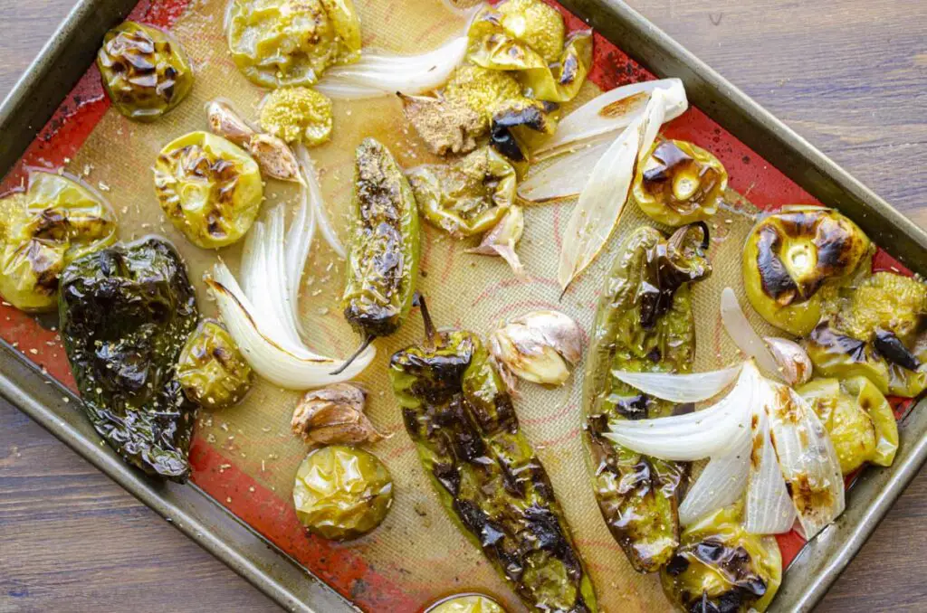 Looking down onto a rimmed baking sheet full of roasted green chiles, tomatillos, onion, garlic and spices.