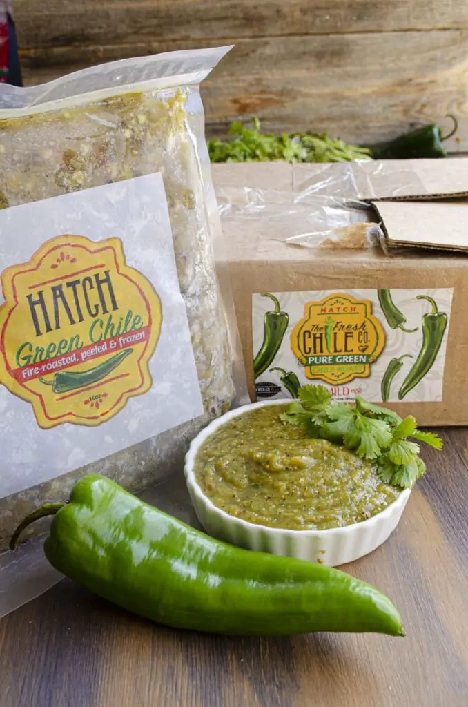 A cardboard box and a 1 pound pouch of frozen Hatch green chile are displayed on a wooden table next to a bowl of Green chili salsa.