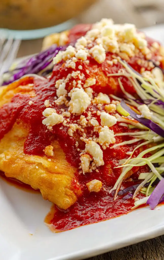 A closeup of stuffed sopapillas smothered in red chile sauce and garnished with fresh cheese and shredded cabbage.