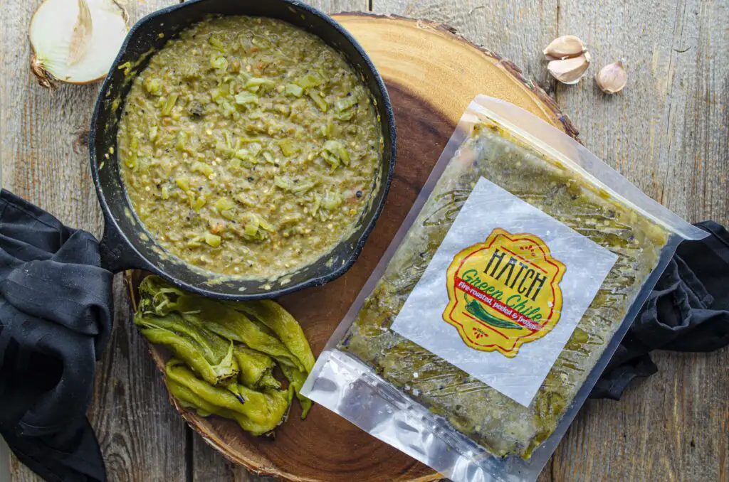 Looking down on a wooden table with a cast iron skillet full of green chile sauce. Next to it is freshly roasted green chile peppers and a package of frozen Hatch green chile.