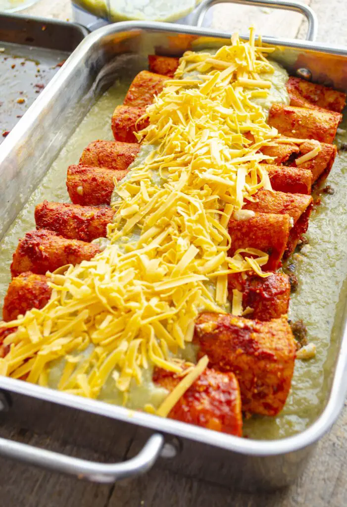 A pan of completely assembled Christmas enchiladas, covered in shredded cheddar cheese, is ready for the oven.
