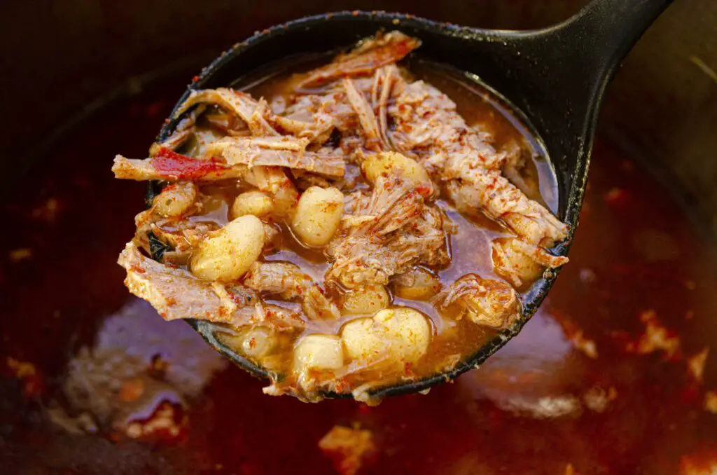 A closeup picture of a soup ladle filled with red chile pork Pozole.