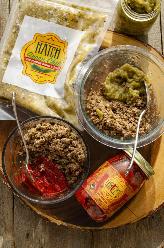 Looking down on a wooden tabletop showing clear glass Bowls filled with ground beef and Hatch red and green chile. A Hatch chile store jar and packet are nestled in between.