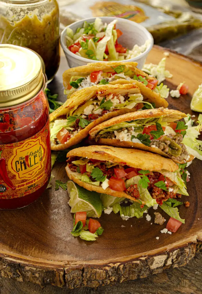 Gorditas filled with ground beef flavored Hatch chile sauce sit on a wooden table. A jar of red chile sauce sits next to the gorditas.