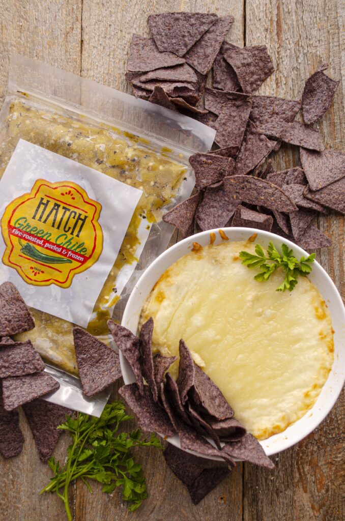 Lookind down on a wooden table with a white ceramic dish full of Skinny Hatch Green Chile Chicken dip surrounded by blue corn tortilla chips and a package of Hatch Green Chile.