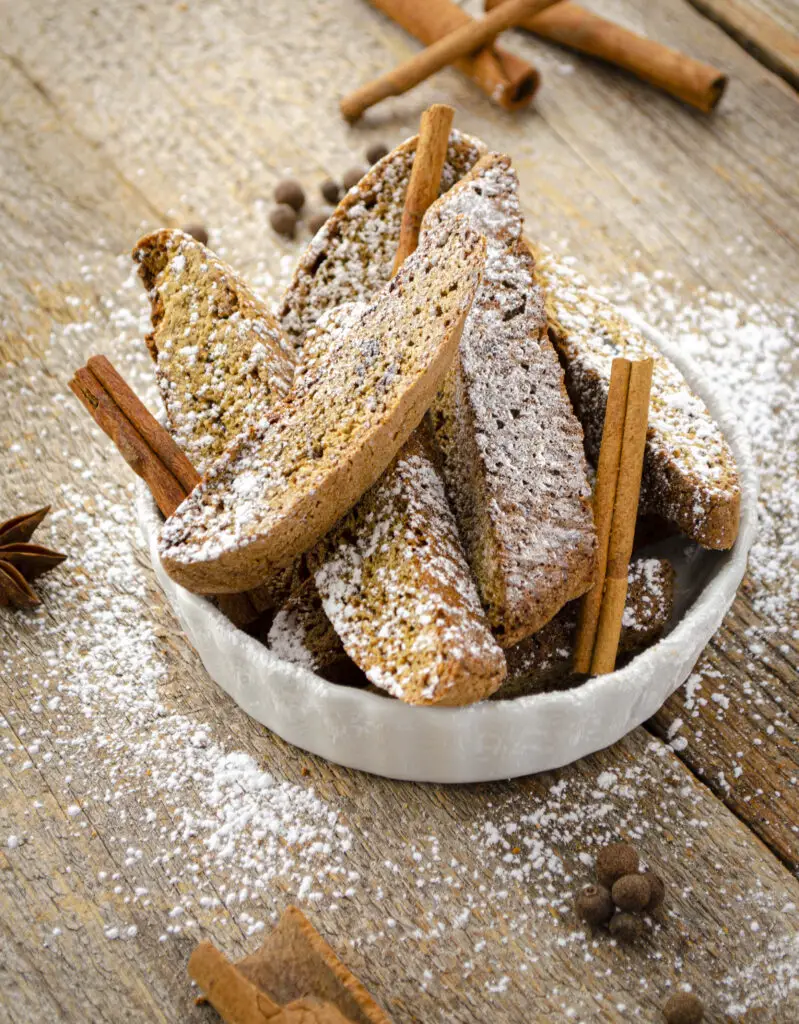 Several brown oval-shapped Christmas Spice Biscotti sit in a white scalloped dish on a wooden tabletop. Powdered sugar is sprinkled over the cookies.
