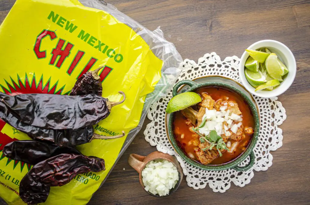 Looking down on a wooden table with a bowl of menudo next to a bag of dried New Mexico red chiles.