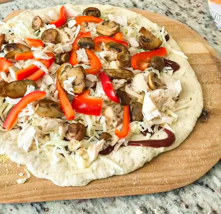 An uncooked BBQ Chicken Pizza made with regular pizza crust dough, topped with BBQ sauce, fully-cooked shredded or chopped BBQ chicken , shredded mozzarella or Monterrey Jack cheese, and toppings such as sliced red onion, bacon, mushrooms, olives, or bell peppers.