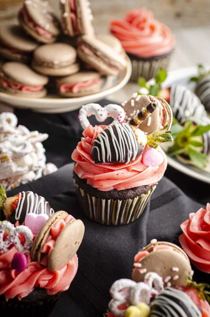 A Dark Chocolate Loaded Valentine's Day Cupcake sits elevated on a black cloth, surrounded by Valentine's day cookies, candies and more cupcakes.