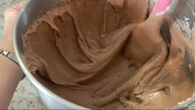A picture of the macaronage process being done on bowl of chocolate batter, with the batter being smeared up the sides of the bowl with a pink spatula to deflate it to the proper consistency. This is macaronage for making macaron cookies.