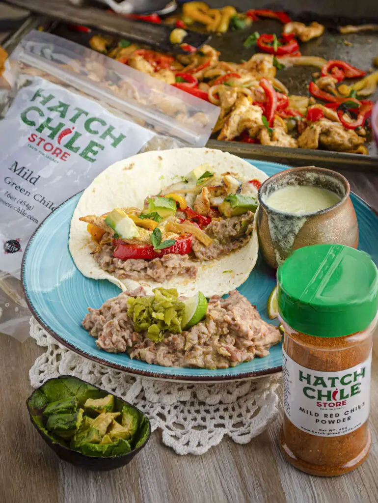 A turquoise dinnerplate features a Sheet Pan Chicken Fajita, with a side of refried beans garnished with limes and chopped Hatch Green chile. The Hatch Chile store products used in the recipe are also displayed.