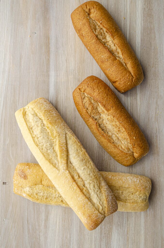 Looking down on a white wooden tabletop spread with french baguettes and wheat sub sandwich rolls.