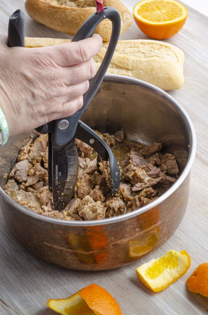 A hand holds shredding scissors in a pot of shredded carnitas.