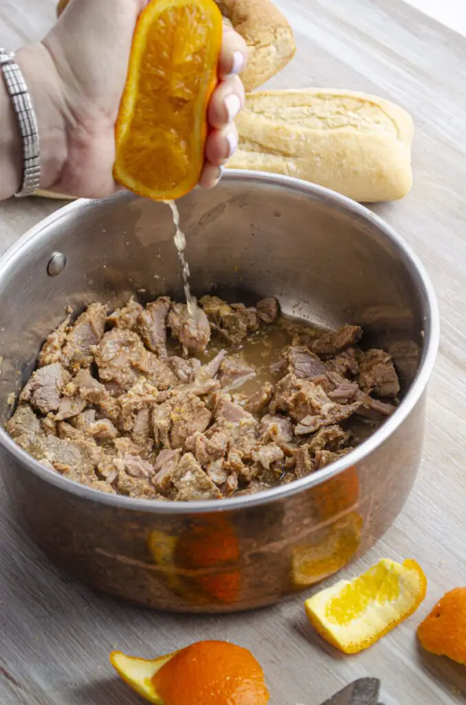Half an orange is squeezed over a pot containing pork carnitas, dripping juice over the meat.
