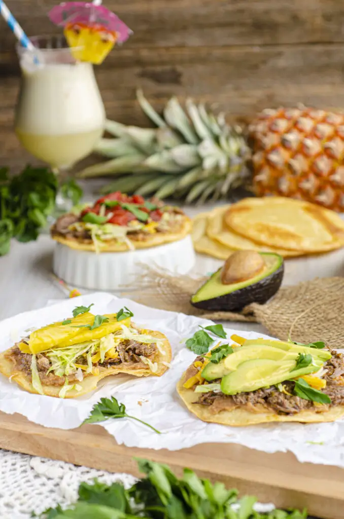 A view of several barbacoa tostadas topped with different toppings such as pineapple, roasted tomato, and avocado. There is a pina colada in the background with an umbrella and a pineapple.