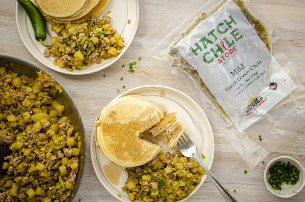Looking down on a white wooden tabletop set with two servings of Hatch green chile breakfast hash, served with stacks of pancakes. A Hatch chile store packet of chile is displayed along side the food.