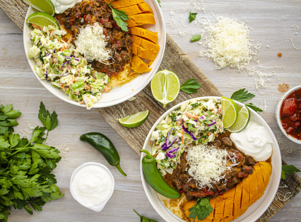 Looking down on a wooden table displaying two white plates each filled with Steak Picada, steamed sweet potato, side salad, and garnishes of sour cream, cheese, and lime wedges.