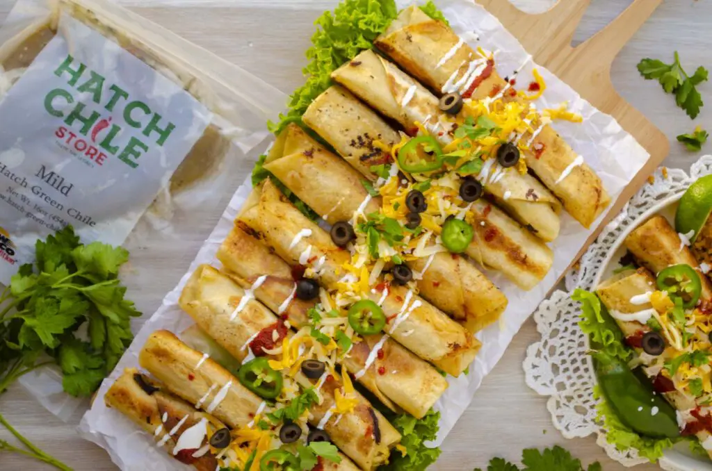 A rectangular wooden platter is filled with Green Chile Chicken Flautas piled with garnishes down the middle. A package of Hatch Chile sits next to the flautas.