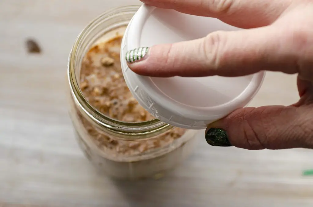 A mason jar of overnight oats made with Naked Oats brand protein powder is shown with a white screw-top lid being placed on it.