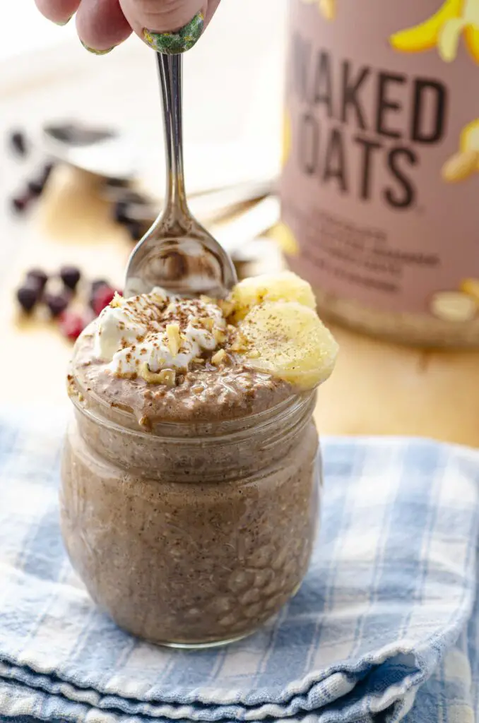 A clear mason jar full to the brim with Chocolate oats is topped with a dollop of yogurt, two slices of banana and a sprinkle of chopped nuts and cocoa powder. A hand is dipping a spoon into the mixture.