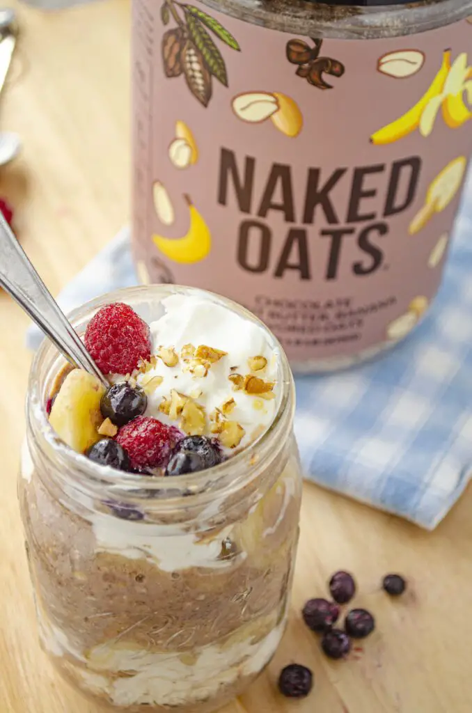 A side view of a mason jar full of overnight oats made with Naked Oats shows it is also mixed with fresh banana slices, yogurt, berries and chopped nuts. A jar of Naked Oats sits beside it.