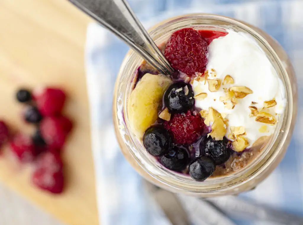 Looking down into a mason jar filled  with overnight chocolate peanut butter banana Naked Oats and topped with yogurt, berries and a sprinkle of chopped nuts. The handle of a spoon is seen coming out of the top of the jar.