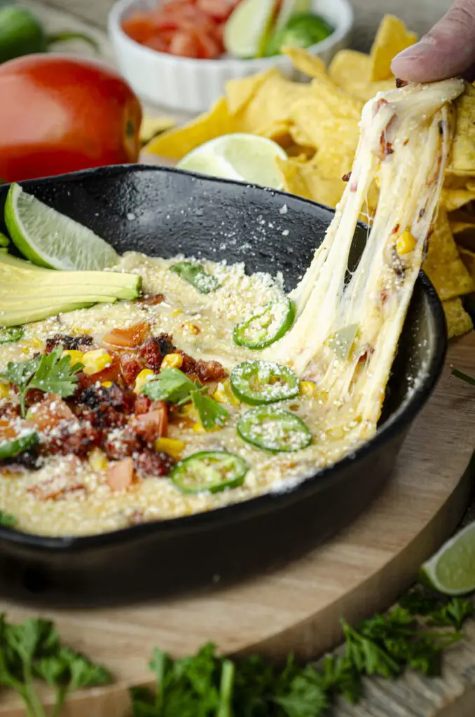 White cheese stretches and pulls as a tortilla chip is dipped in queso fundido, garnished with sliced jalapeno, chorizo, siced jalapeno, corn, and tomato.