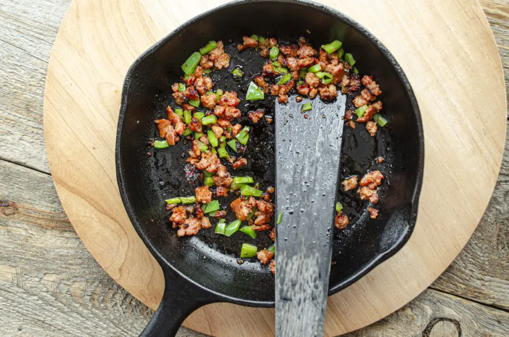 Looking down into a cast-iron skillet where crumbled chorizo sausage and chopped green chile are cooking.